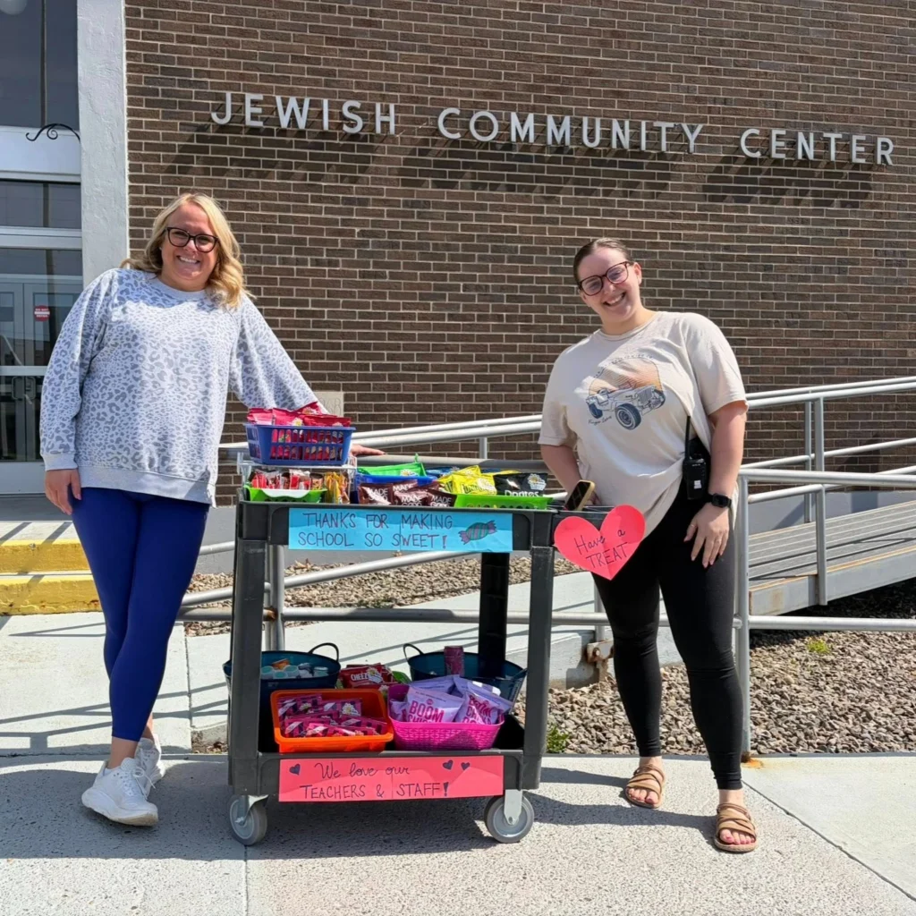 Two women stand beside an Auchinachie Cares snack cart with thank-you signs in front of a Jewish Community Center building.