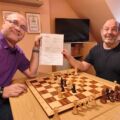 Zwei Männer spielen Schach, einer übergibt einen Preis, im Hintergrund Fernseher.