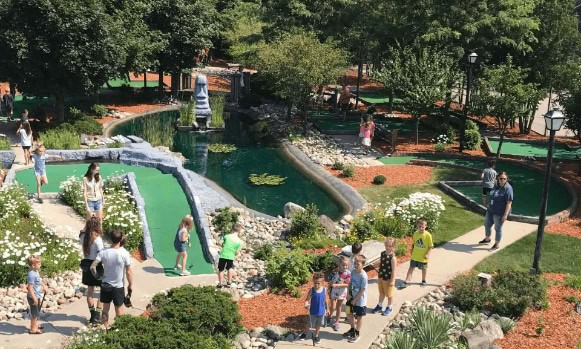 Colorful park scene with children playing near water features and families walking along scenic path.