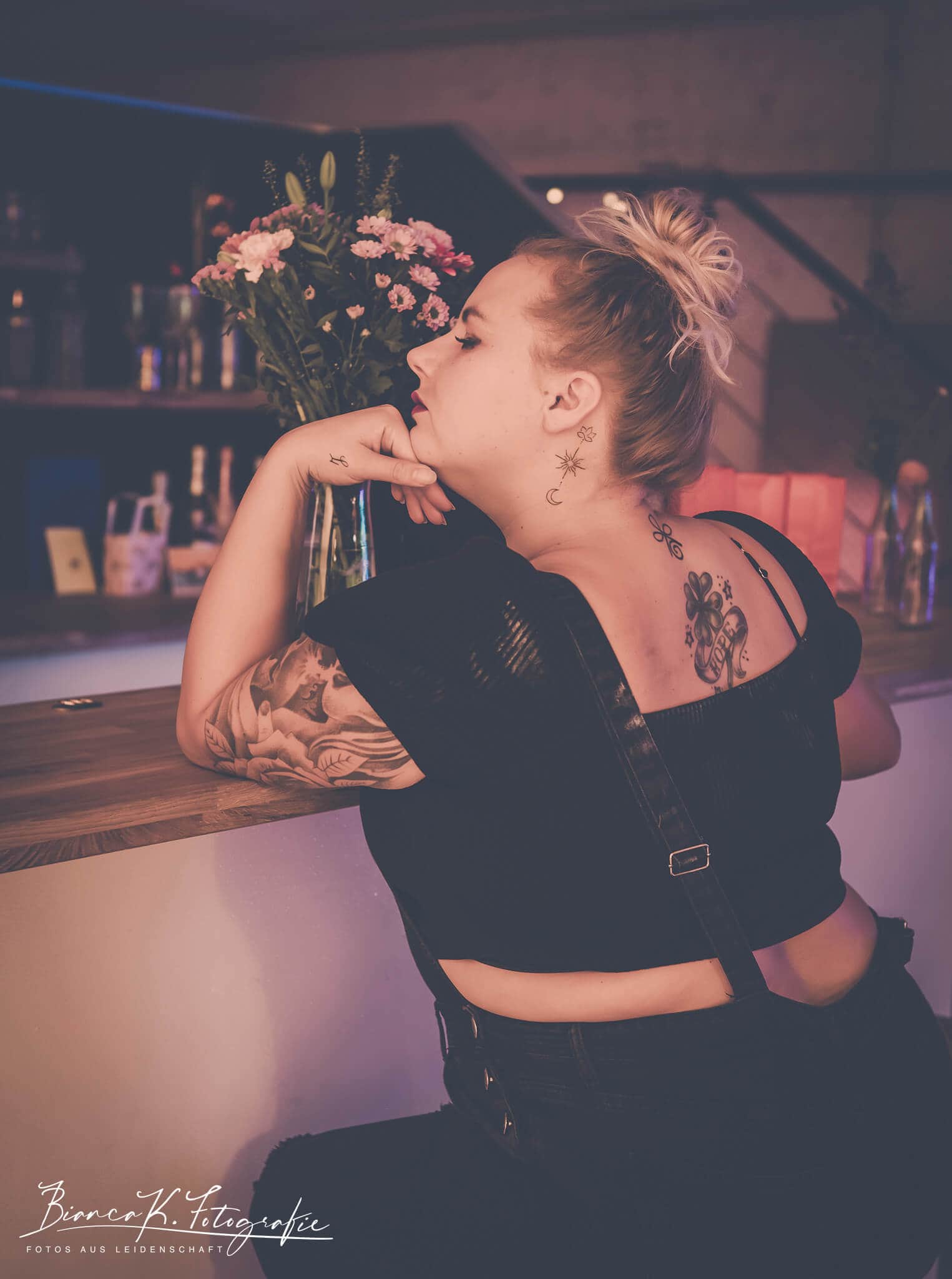 Moderne Portraitaufnahme einer Tattoo-Trägerin in entspannter Atmosphäre in Aachen. Stilvolle tattoos einer jungen Frau in einem bar-ambiente, abendliche Atmosphäre, kreative Portraitaufnahme, Graffiti- und Bar-Hintergrund, moderne Tattoo-Kunst.