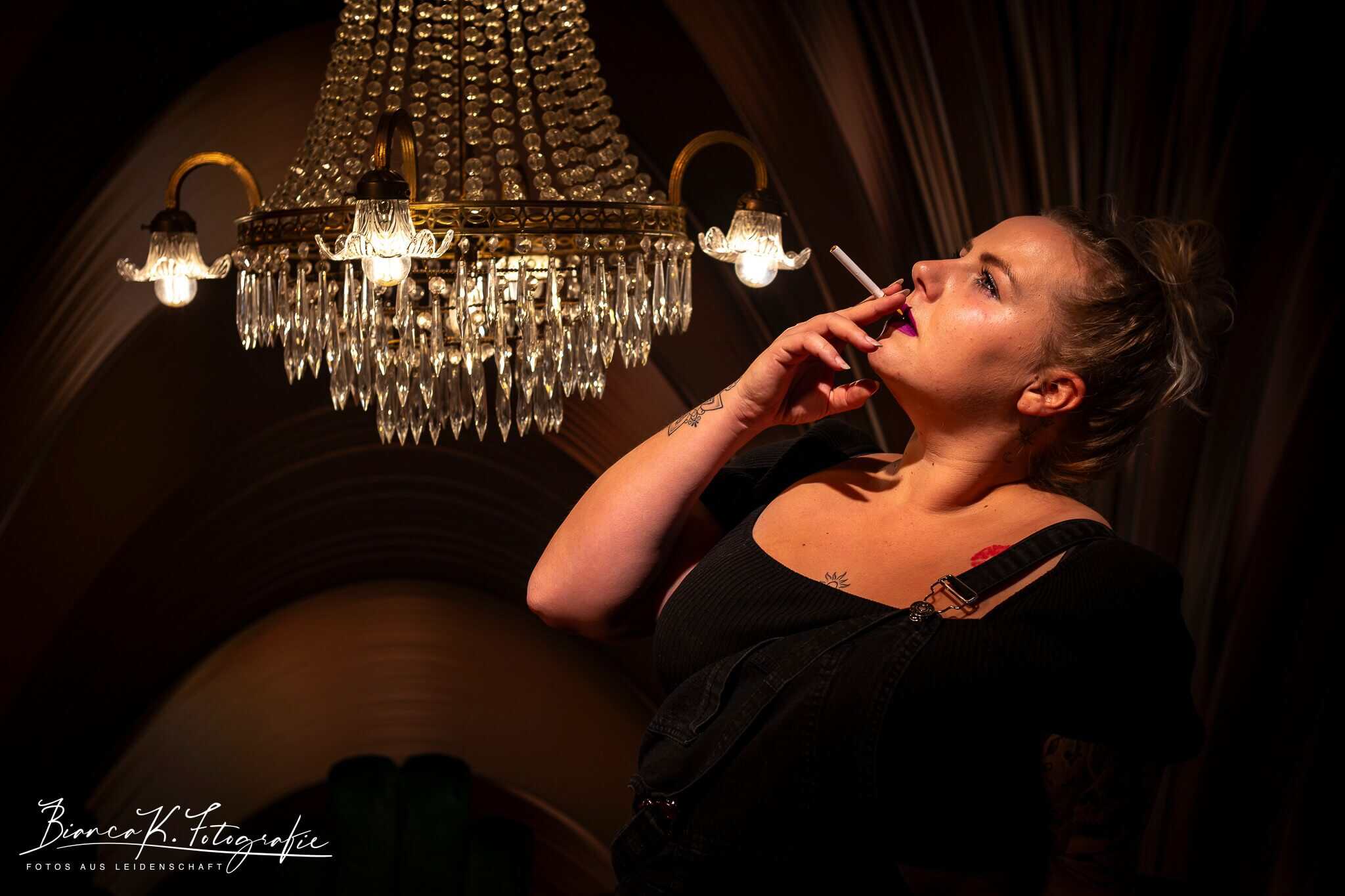 Aufgenommen bei einem Fotoshooting in Aachen mit stilvoller Beleuchtung und urbanem Ambiente. Glückliche junge Frau raucht und stärkt sich in einem eleganten Raum mit Kronleuchter.