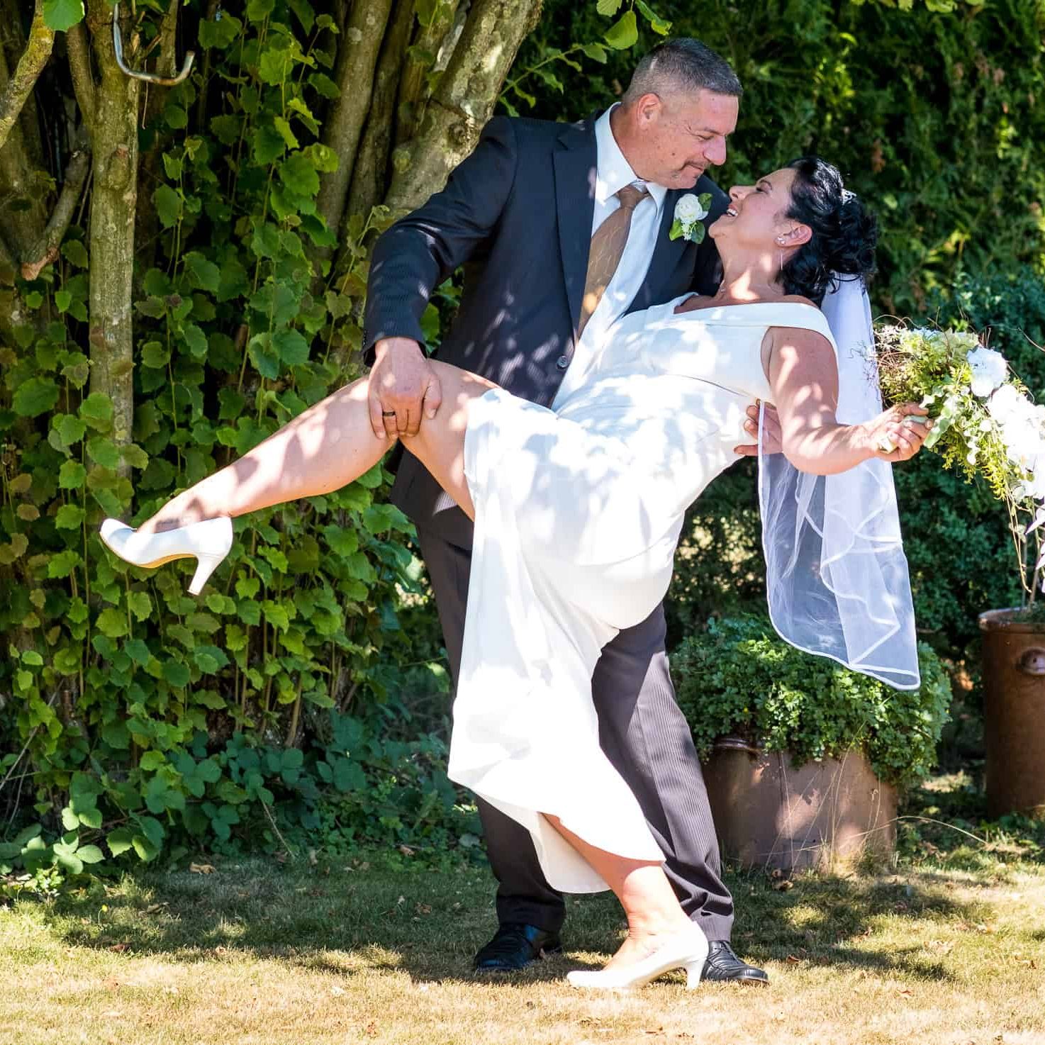 Hochzeitspaar im Garten, Braut in weißem Kleid mit Blumenstrauß, langer Schleier, lachendes Paar.