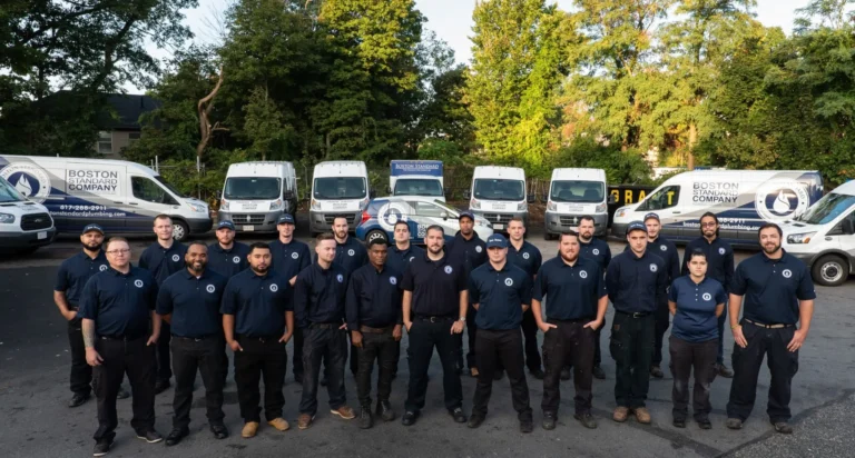 A team of 19 people in navy uniforms stands proudly in front of a line of company vans with "Boston Standard Company" logos, set against a backdrop of trees, embodying the spirit of rewarding careers and exciting job opportunities.