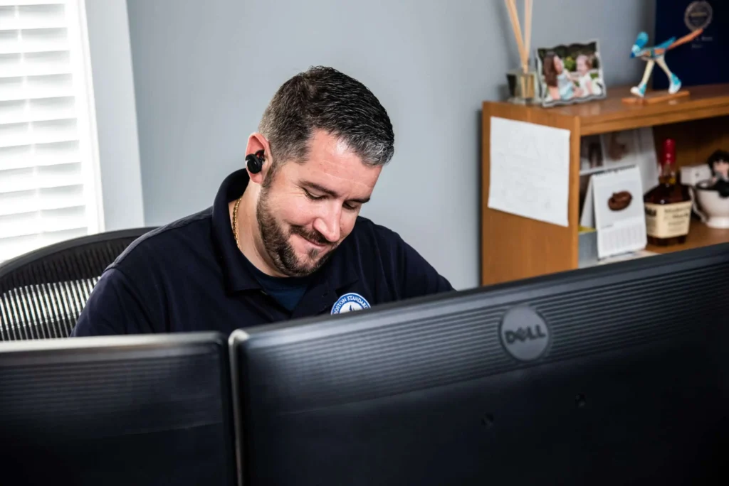 Man with earbuds smiling, sitting at a desk with dual Dell monitors, a bookshelf with photos and a bottle—immersed in his work, as he navigates his career in this well-lit room.