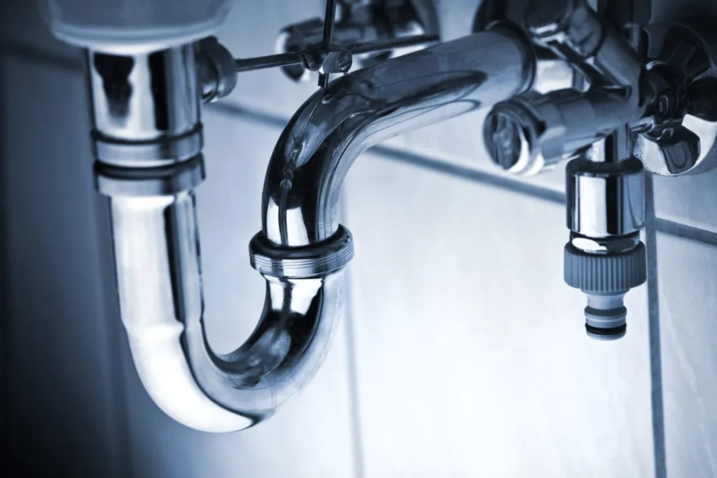 Close-up of a chrome sink drain pipe with visible fittings against a tiled wall.