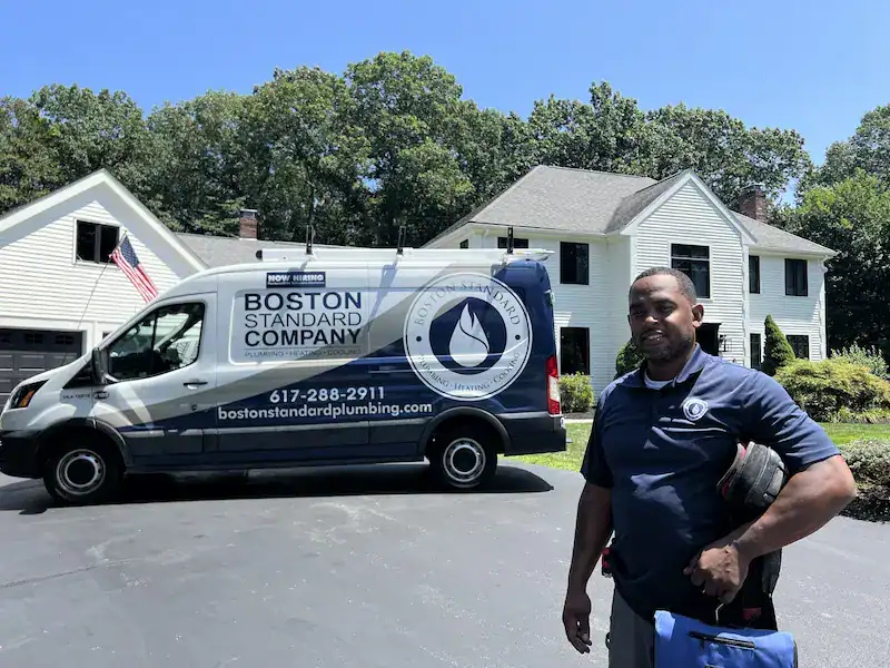 A Boston Standard Company plumber stands in front of a company van, showcasing their expertise as part of the Premier Pro Program, parked outside a two-story suburban house on a sunny day.