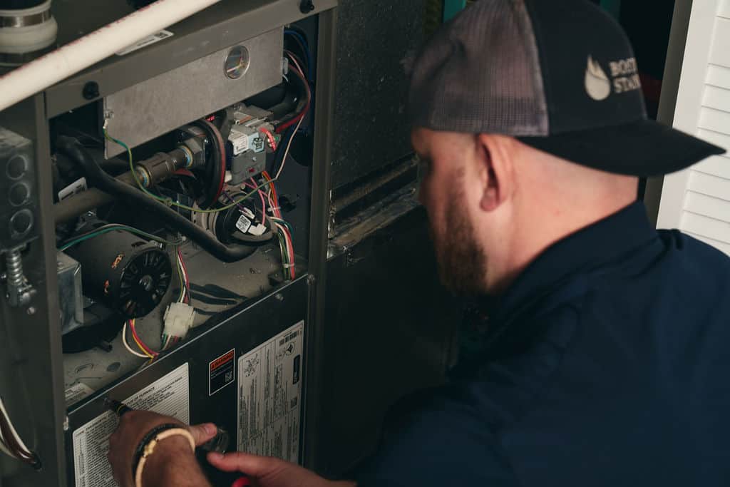 A technician wearing a cap repairs or inspects the inside of a furnace or HVAC unit, using tools and examining wiring and components—ensuring everything is ready for HVAC upgrades and SEER2 standards.