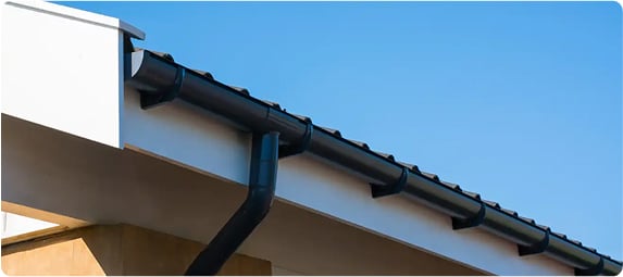 Close-up of a newly installed black gutter system along the edge of a roofline under clear blue skies.