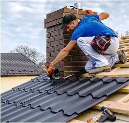 A roofing professional attaching panels of dark metal roofing around a brick chimney on a sloped roof.