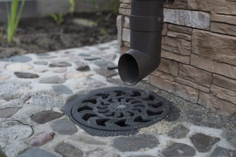 A black downspout directs water toward a round, decorative drain grate set in a stone-paved surface next to a stone wall.