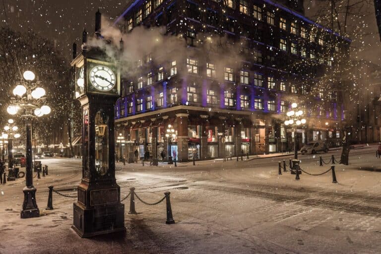 A steam clock stands on a snowy city street at night, with illuminated buildings and streetlights in the background as snow falls.
