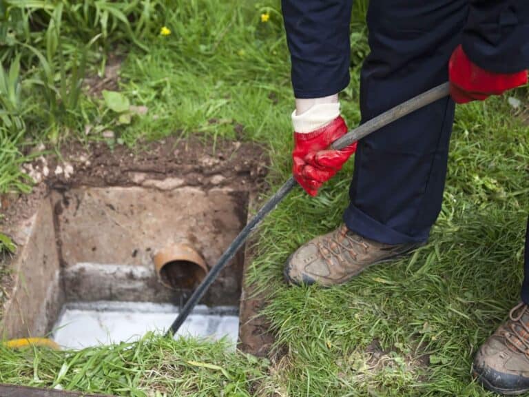 Person wearing gloves and work boots using a metal rod to clean or inspect a drain or sewer pipe in a grassy outdoor area.