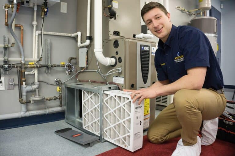 A technician kneels next to an HVAC unit, preparing to replace an air filter, with tools and pipes visible in the background.