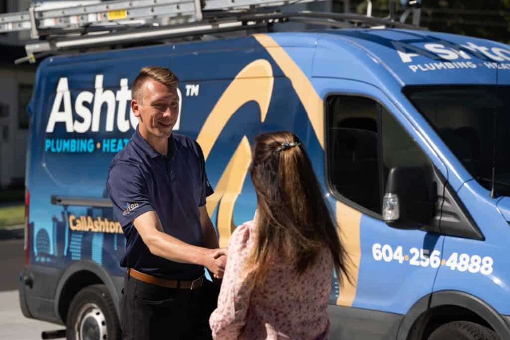 A man in a blue uniform shakes hands with a woman in front of a blue Ashton Plumbing service van.