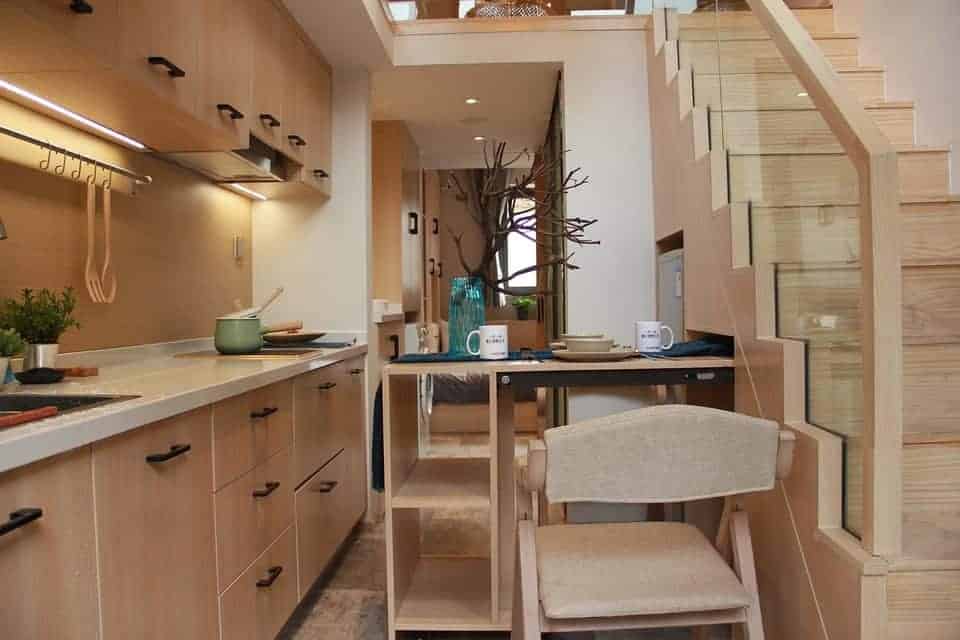 Modern compact kitchen with light wood cabinets, small dining table set for two, and staircase with glass railing leading to an upper level.