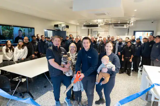 A group of people pose indoors at a ribbon-cutting event; one person holds large scissors, another holds a dog, and one holds a baby.