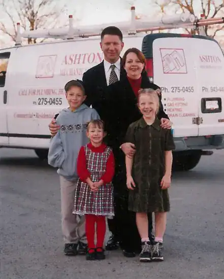 A family of five poses together in a parking lot in front of a white plumbing service van labeled “Ashton Mechanical.”.