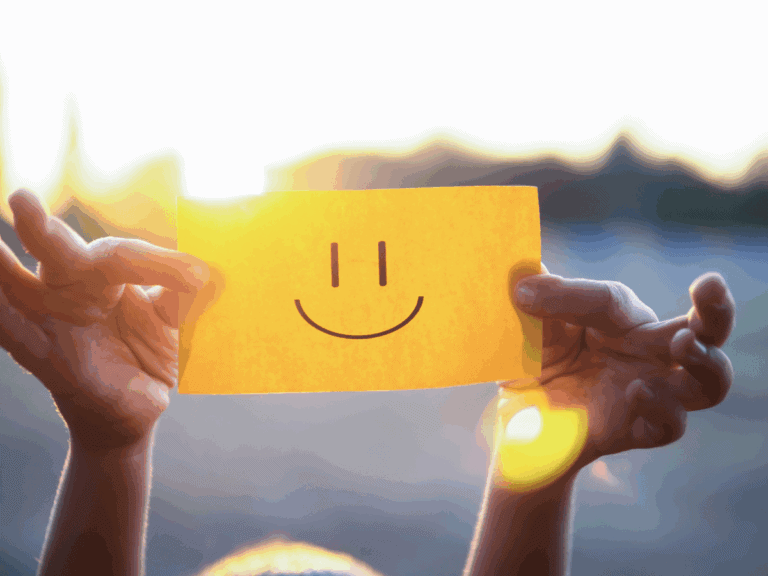 A person holds up a yellow paper with a simple black smiley face drawn on it, with sunlight shining in the background.