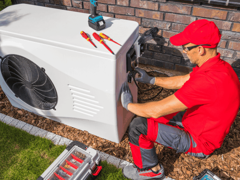 A technician in red uniform repairs a white outdoor heat pump unit, using tools laid out on top and beside the unit.