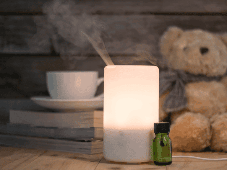 A white essential oil diffuser emits vapor beside a green bottle, stacked books, a cup and saucer, and a teddy bear on a wooden surface.
