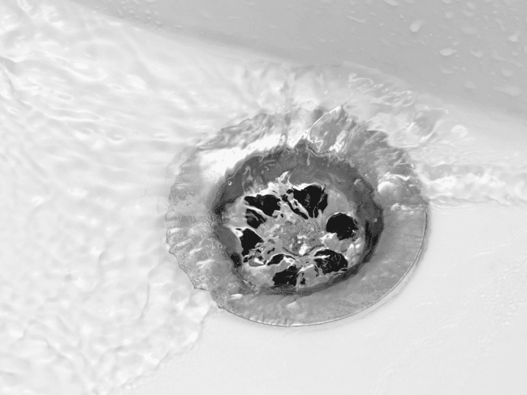 Water flows toward a metal drain in a white sink, creating ripples and swirling patterns around the drain cover.