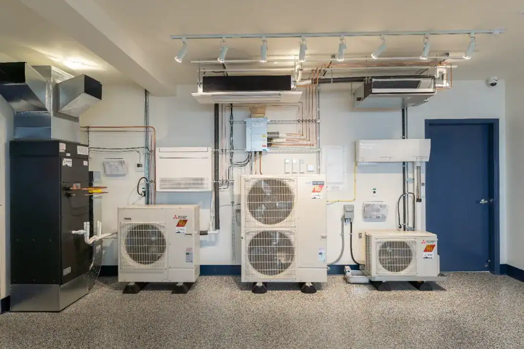 Three indoor HVAC units and connected ductwork are mounted on a wall in a utility room with a blue door and speckled flooring.