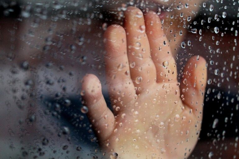A child's hand touches a rain-covered window, with water droplets scattered across the glass.