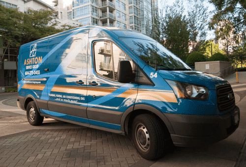 Blue Ashtion service van parked on a paved area in front of modern apartment buildings, with trees and sunlight in the background.