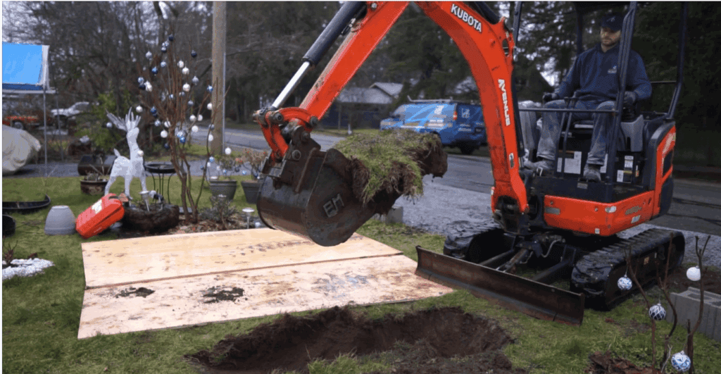 A person operates an orange mini excavator to dig a hole and lift a chunk of grass and soil from a residential lawn, with plywood sheets protecting the ground nearby.
