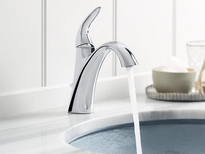 A modern chrome bathroom faucet with water running into a sink, set on a white countertop with a soap dish and glass in the background.