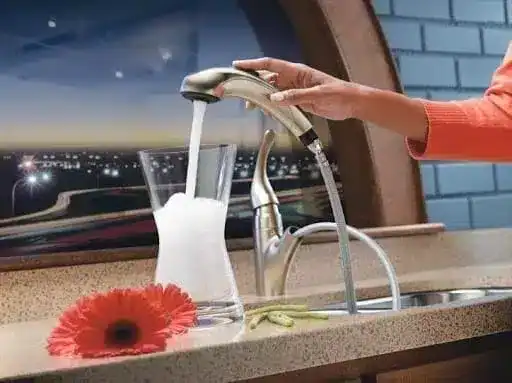 A person fills a clear glass vase with water from a kitchen faucet; a red flower lies on the countertop beside the sink.