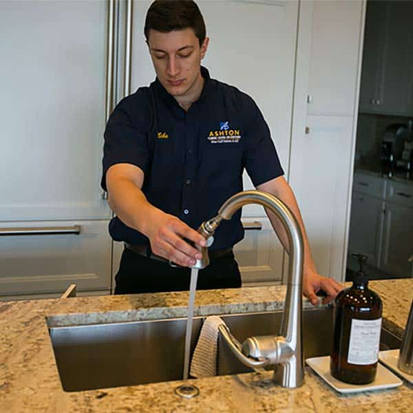A man in a uniform runs water into a kitchen sink, holding the faucet with one hand. A brown bottle and a white sponge are on the counter beside him.