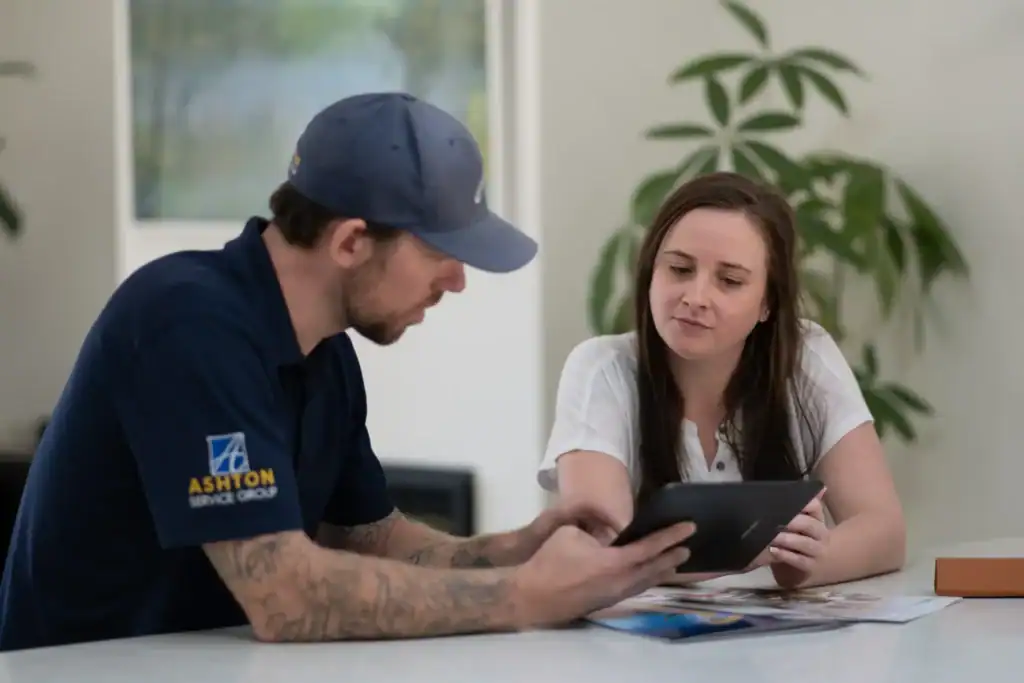 A man in a blue Ashton Service Group shirt shows a tablet to a woman sitting across a table with papers and pamphlets.