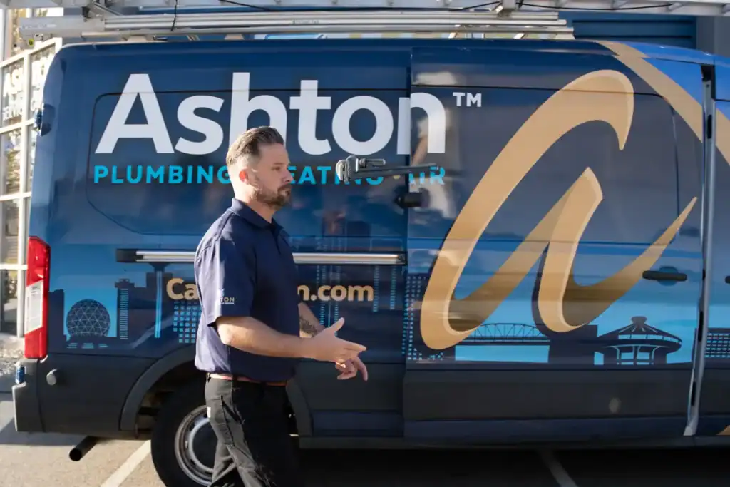 A man in a navy shirt walks past a blue Ashton Plumbing, Heating, and Air van in a parking lot on a sunny day.