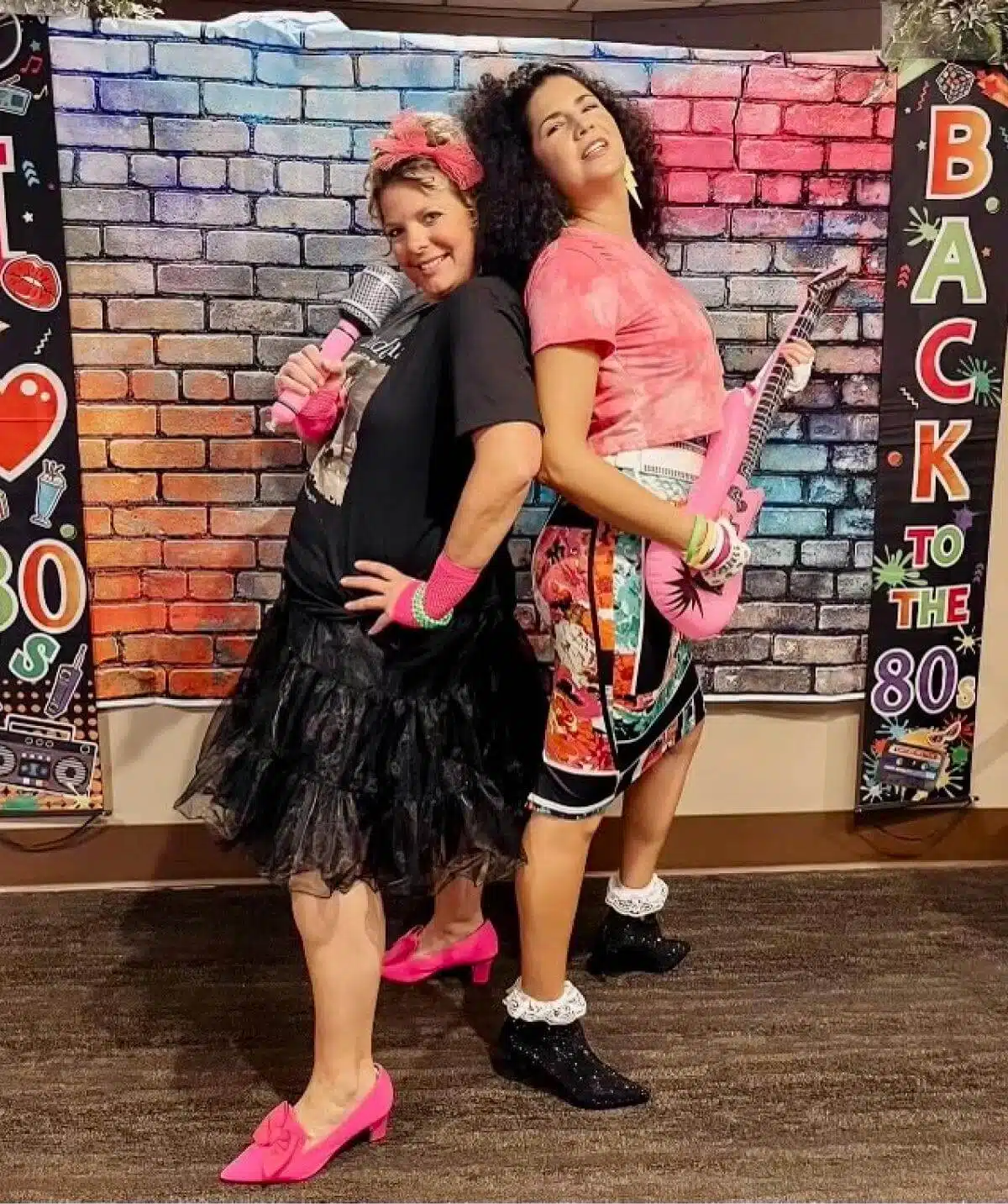 Two women in 80s-themed costumes posing with guitars at Cascades Manor event.