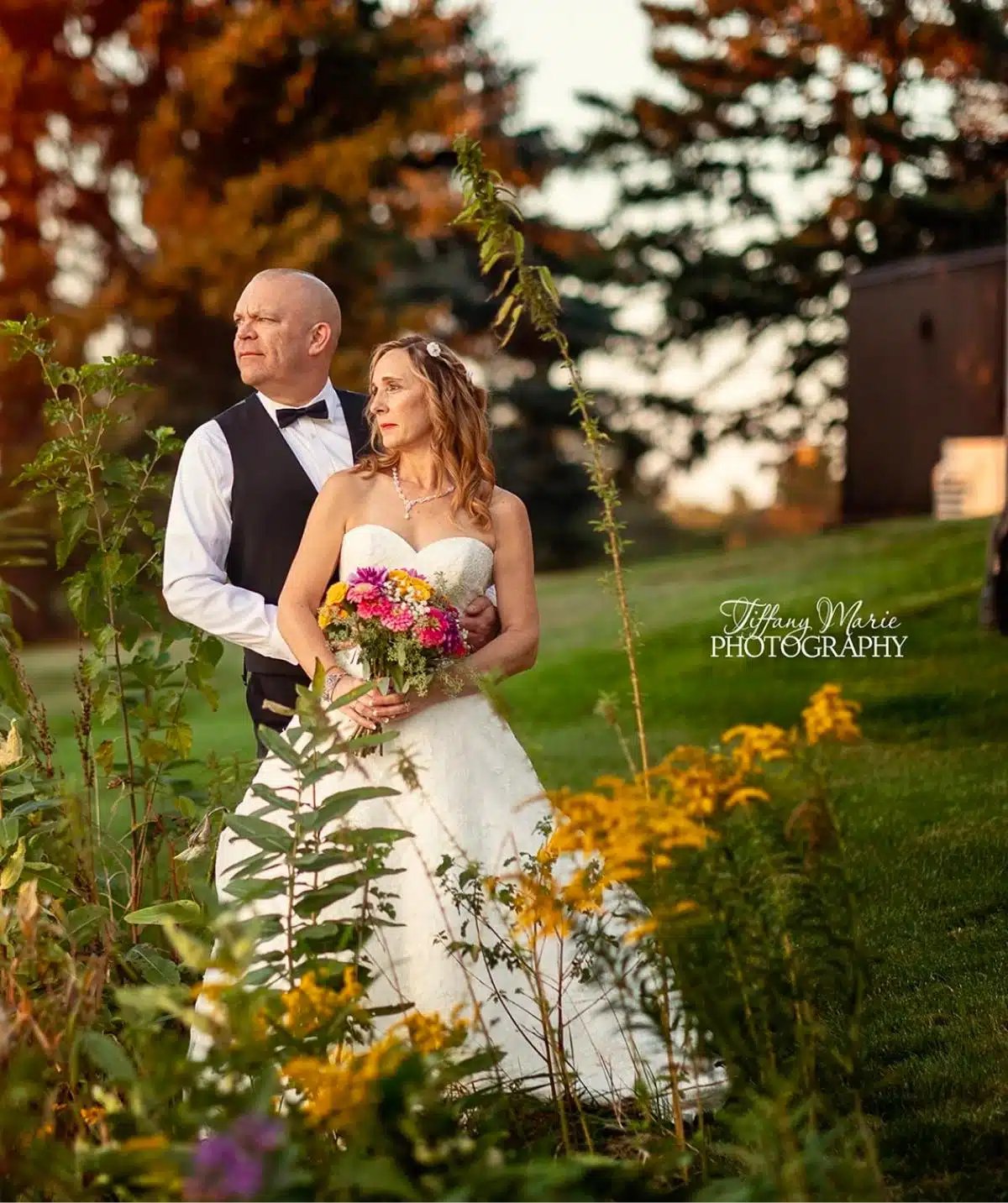 Bride and groom standing outdoors surrounded by colorful flowers and lush greenery.