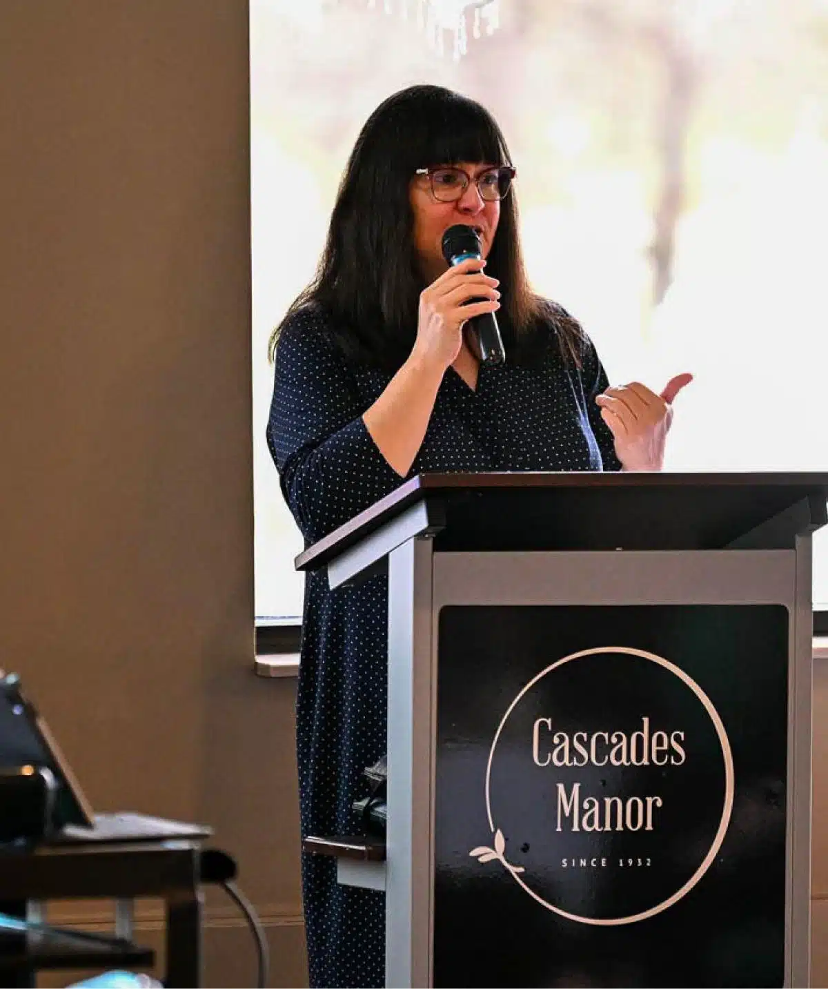 Woman speaking at Cascades Manor event with microphone and podium.