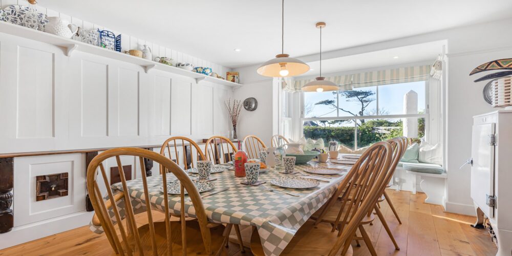 Bright, cosy coastal dining room with wooden furniture, white walls, and large windows overlooking gardens.