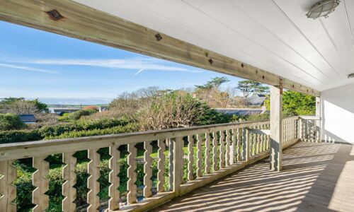 Stunning seaside view from a wooden balcony overlooking lush green gardens and blue skies.