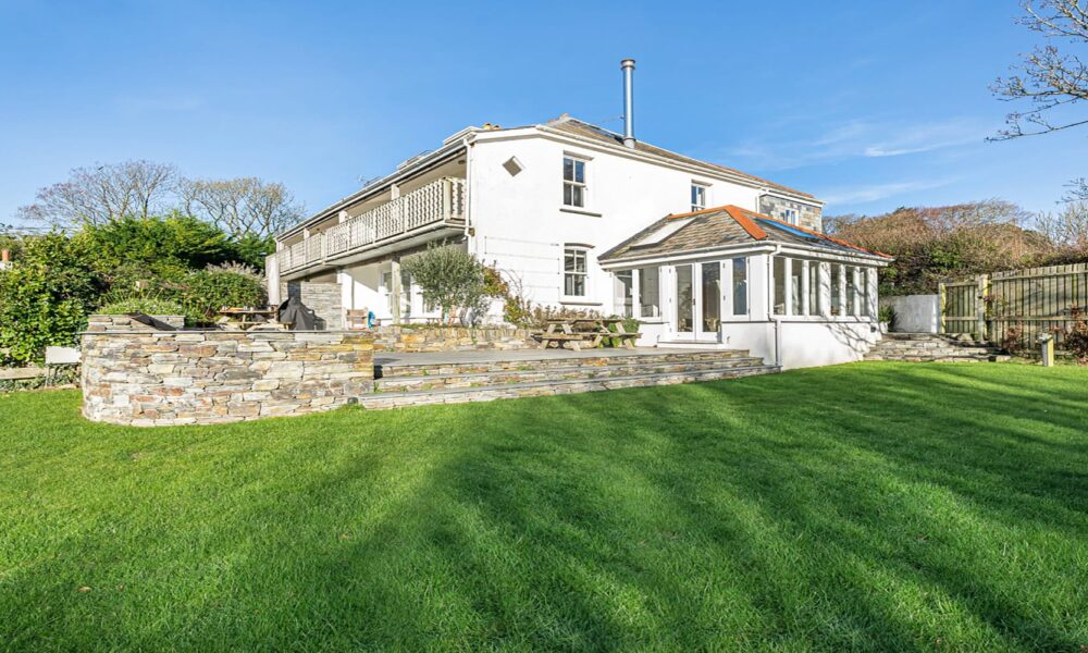 Bright sunny view of a charming Cornish seaside cottage with lush garden and outdoor seating area.