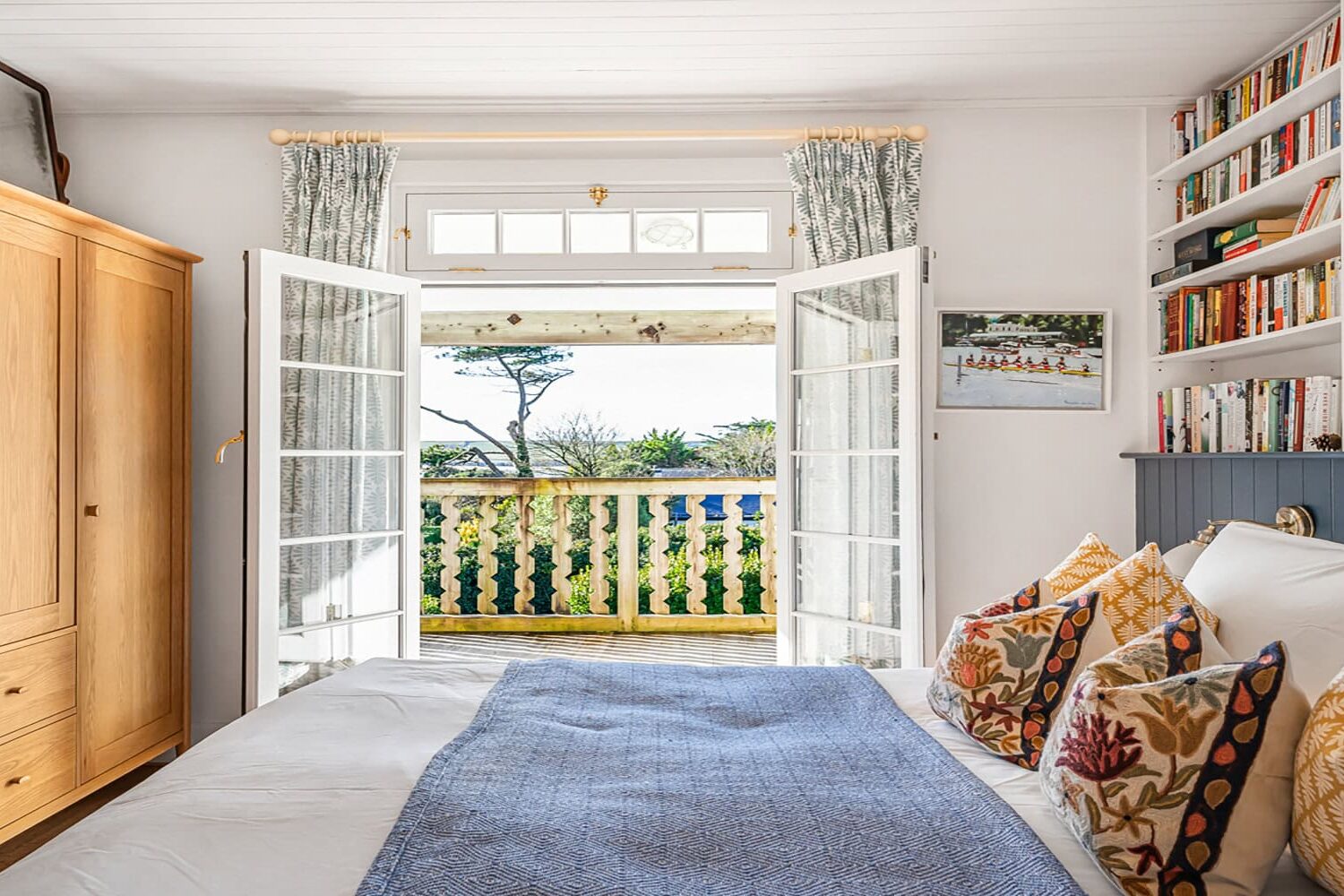 Coastal bedroom with sea view, wooden furniture, colourful cushions, and open French doors to a balcony.