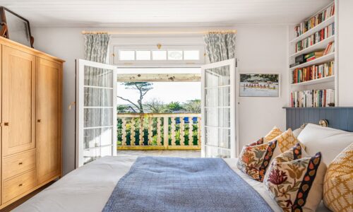 Coastal bedroom with sea view, wooden furniture, colourful cushions, and open French doors to a balcony.