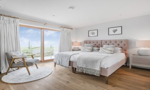 Coastal bedroom with pink upholstered bed, wooden flooring and sea view through large glass doors.