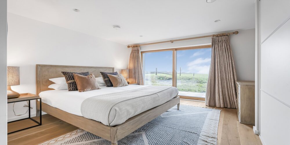 Stylish bedroom with large window overlooking scenic Cornish countryside, warm neutral tones.