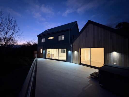 Coastal modern holiday home in Cornwall, illuminated at dusk with a spacious deck area.