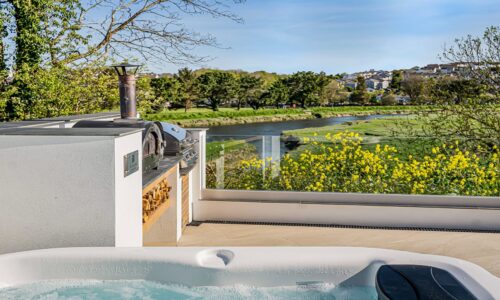 Hot tub overlooking a river with lush greenery and yellow wildflowers in Cornwall.