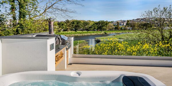 Hot tub overlooking a river with lush greenery and yellow wildflowers in Cornwall.