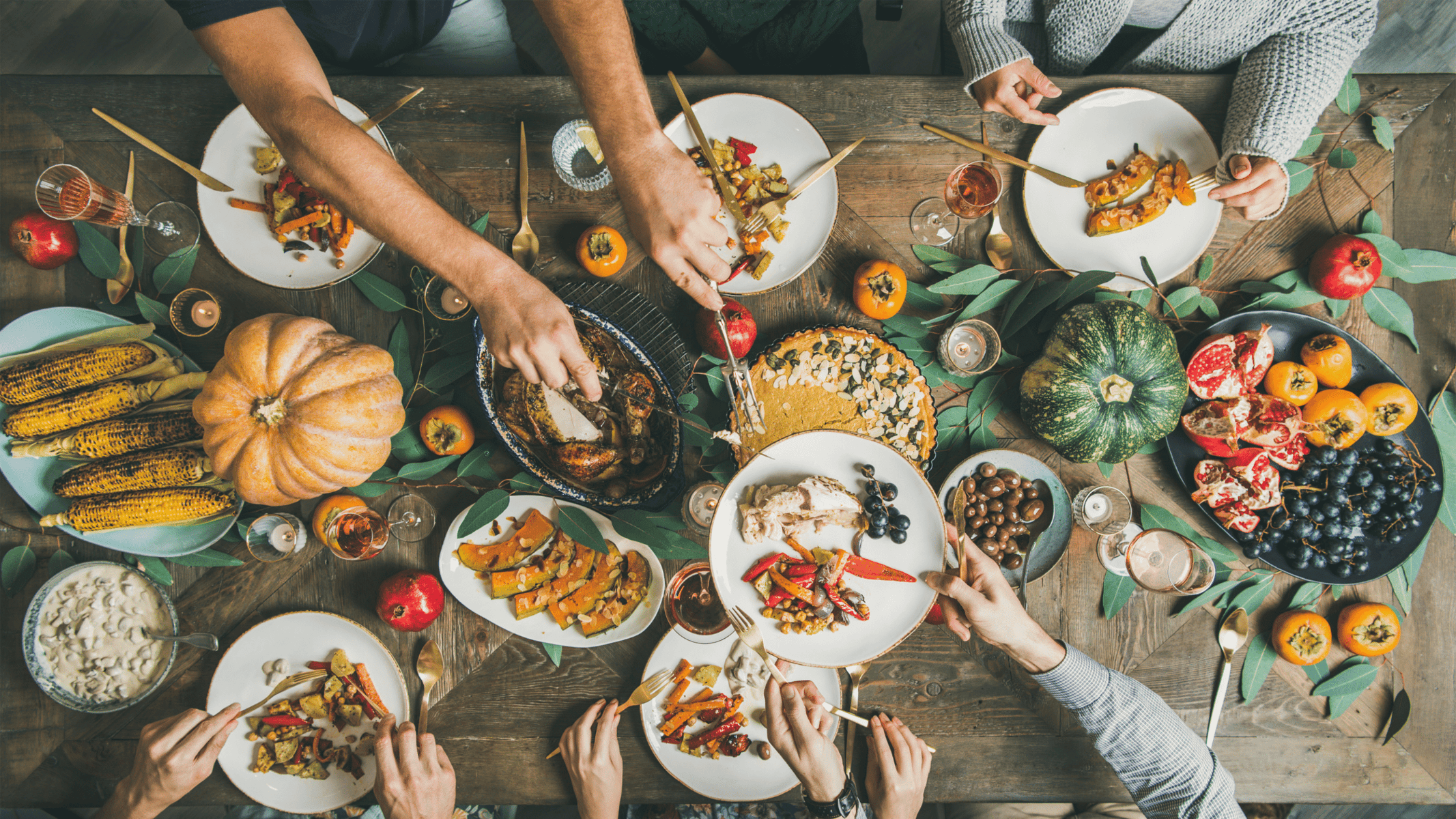Aerial view of a wooden table with people sharing a variety of autumn foods, including pumpkins, roasted vegetables, and fruits. Hands reach for dishes; leaves decorate the table.