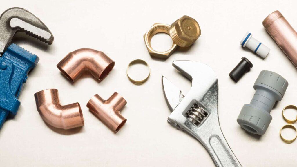A variety of plumbing tools and fittings, including a wrench, copper elbows, connectors, and nuts, are arranged on a white surface.