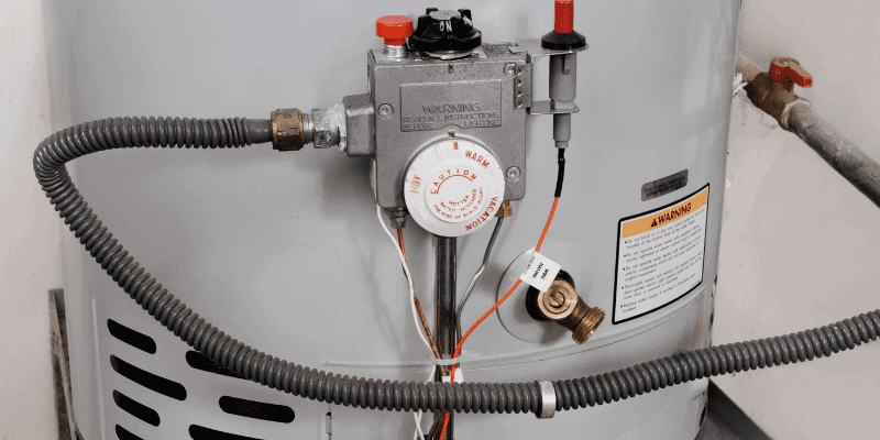 Close-up of a water heater's control panel featuring temperature control dial, warning labels, and wiring, in a utility room.